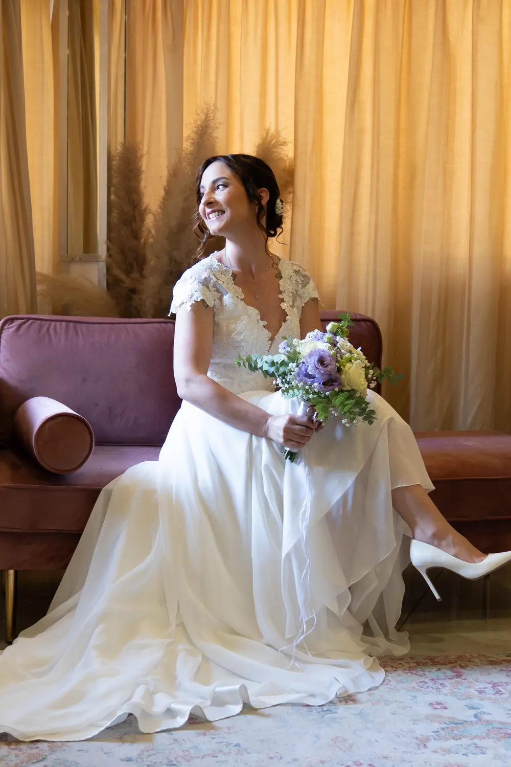 Sposa sorridente su divano, tende dorate, saletta preparazione